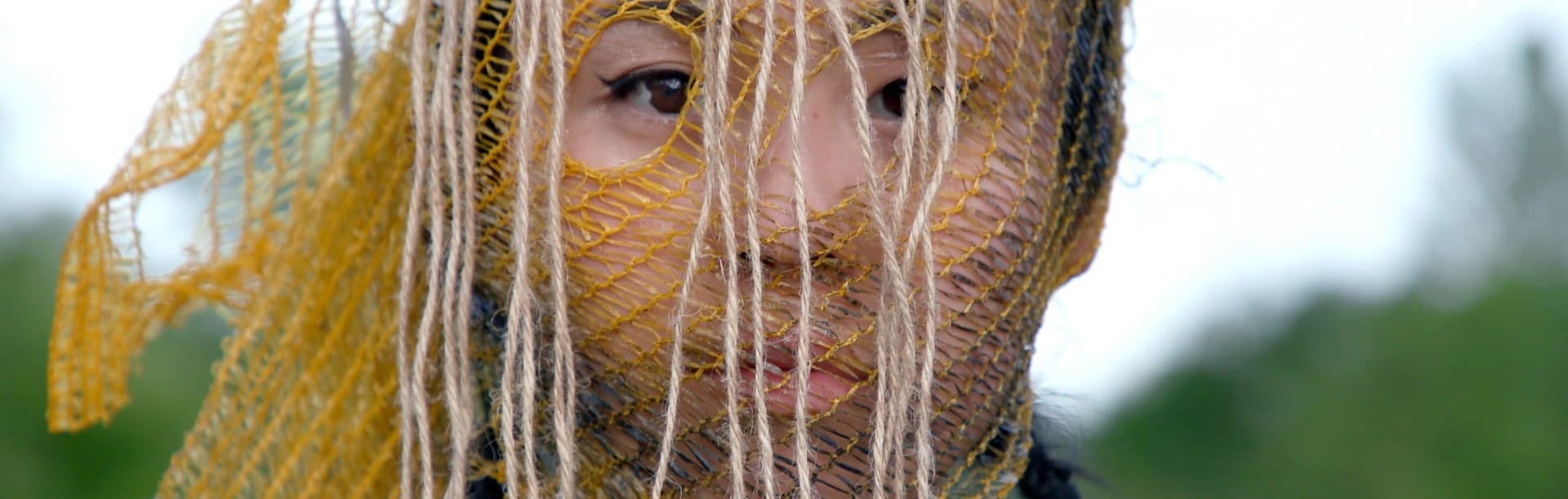 A face behind a yellow mesh and hanging threads, in a field